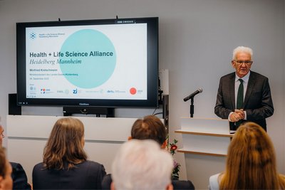 Ministerpräsident Winfried Kretschmann steht hinter einem Stehpult und spricht zu den anwesenden Gästen. Neben ihm ist auf einem großen Bildschirm das Logo der Health + Life Science Alliance Heidelberg Mannheim zu sehen. 