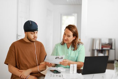 A young man wears a cap with electrodes on his head and is instructed by a practitioner in the use of transcranial direct current stimulation.