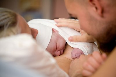 Eine Frau hat ihr Neugeborenes auf dem Bauch und ihr Partner legt die Hand auf das Neugeborene.