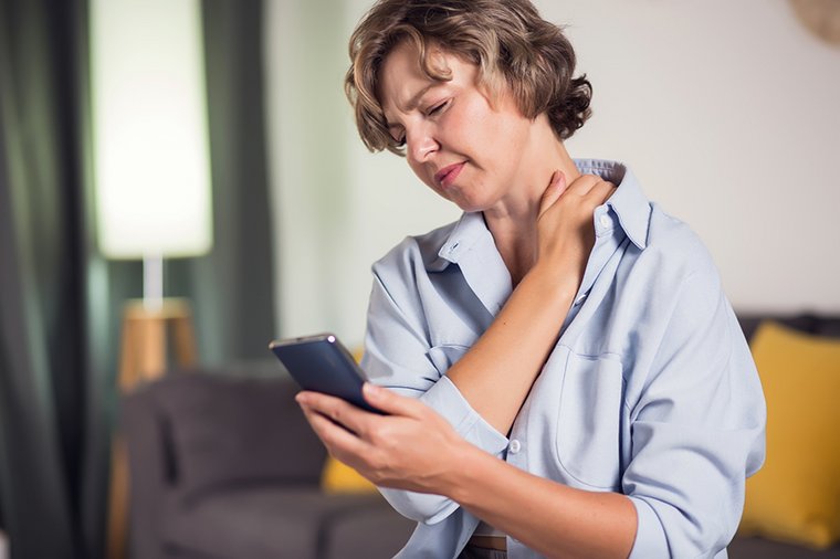 Eine Frau hat offensichtlich Rückenschmerzen. Sie schaut auf ihr Smartphone. 
