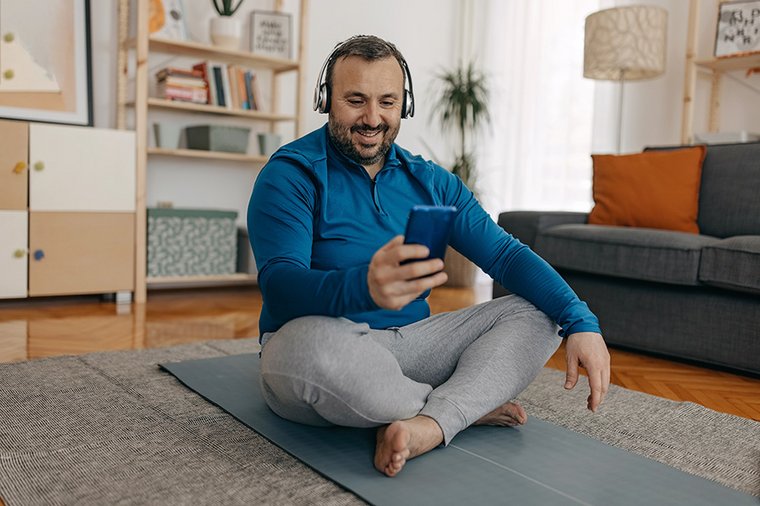 Ein Mann im mittleren Alter sitzt auf einer Sportmatte. Er lächelt und hat ein Smartphone in der Hand. 