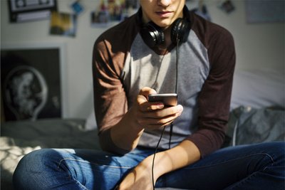 Ein Jugendlicher sitzt in seinem Zimmer auf dem Bett und nutzt sein Smartphone.