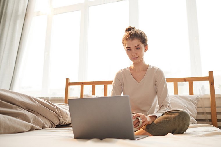 Eine junge Frau sitzt mit einem Laptop auf Ihrem Bett.