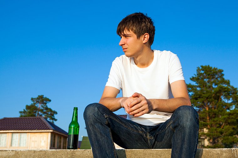 Ein junger Mann sitzt nachdenklich auf einer Mauer, neben ihm eine Bierflasche. 