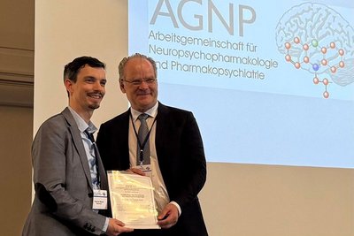 Prof Dr. Dr. Patrick Bach empfängt den Preis auf einer Bühne aus den Händen von Prof. Dr. Matthias Riemenschneider (1. Vorsitzender der AGNP). 
