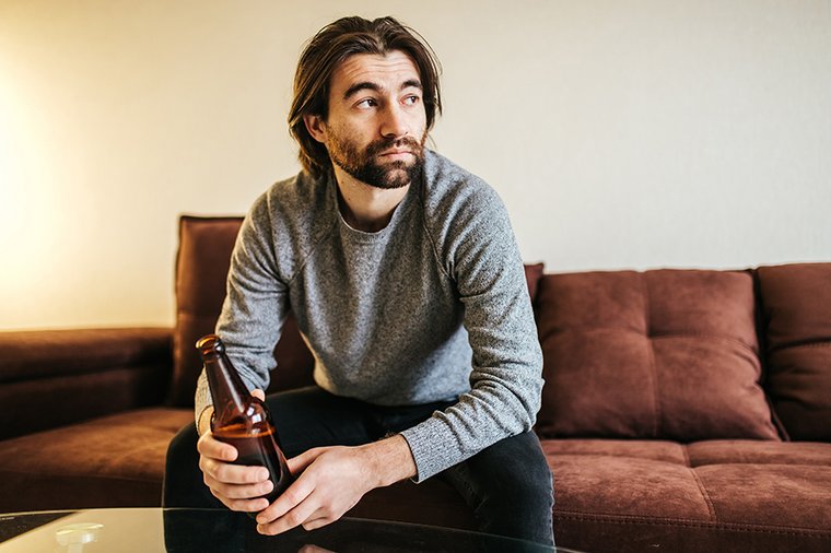 Ein einsam wirkender Mann sitzt mit einer Flasche Bier auf dem Sofa. 