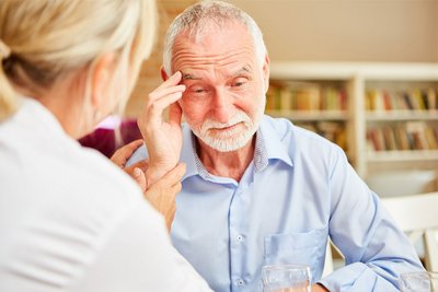 Ein älterer, an Alzheimer erkrankter Mann fasst sich an die Schläfe während er von einer Ärztin Zuspruch erfährt.