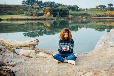 Eine junge Frau sitzt an einem See und schaut in ihr Smartphone. 